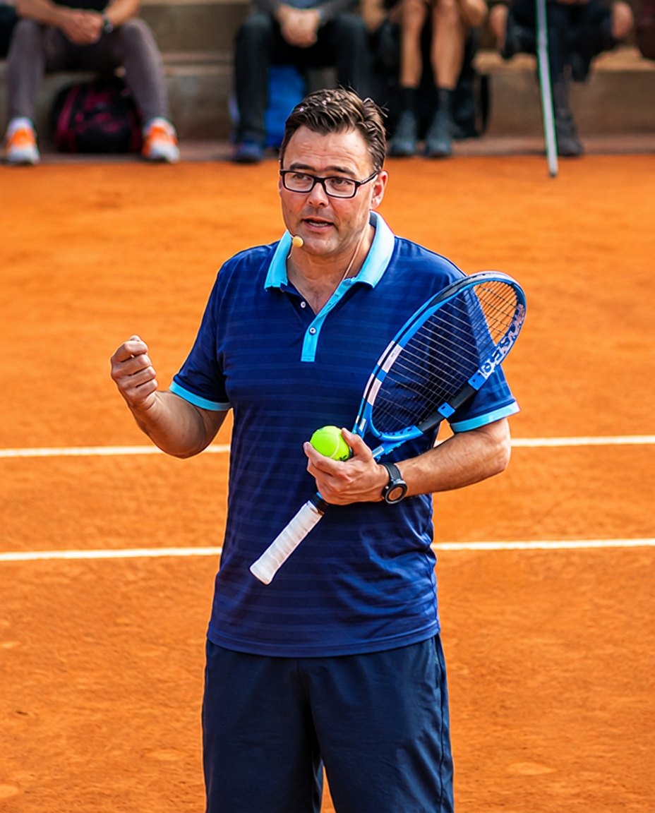 Carl Maes coaching on court, microphone on, racket and ball in hand