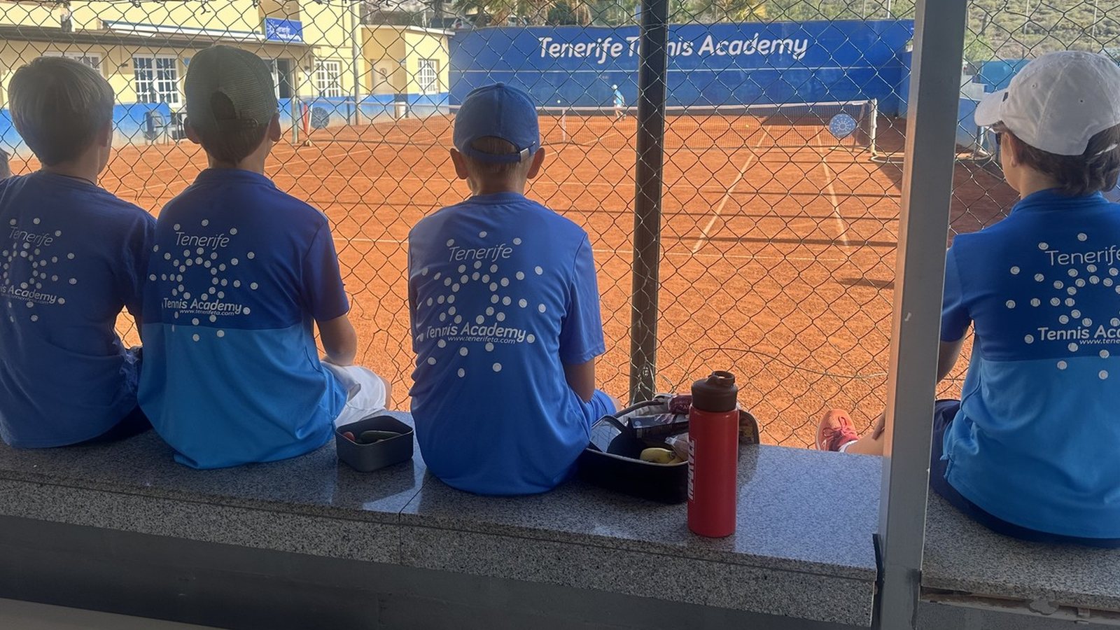 Children in academy shirts sitting on the wall watching a match on the clay at Chayofa