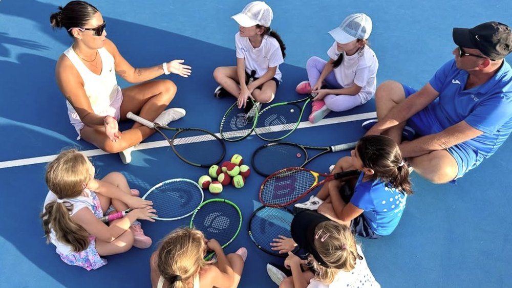 A coach sitting on court with a group of young players and their rackets