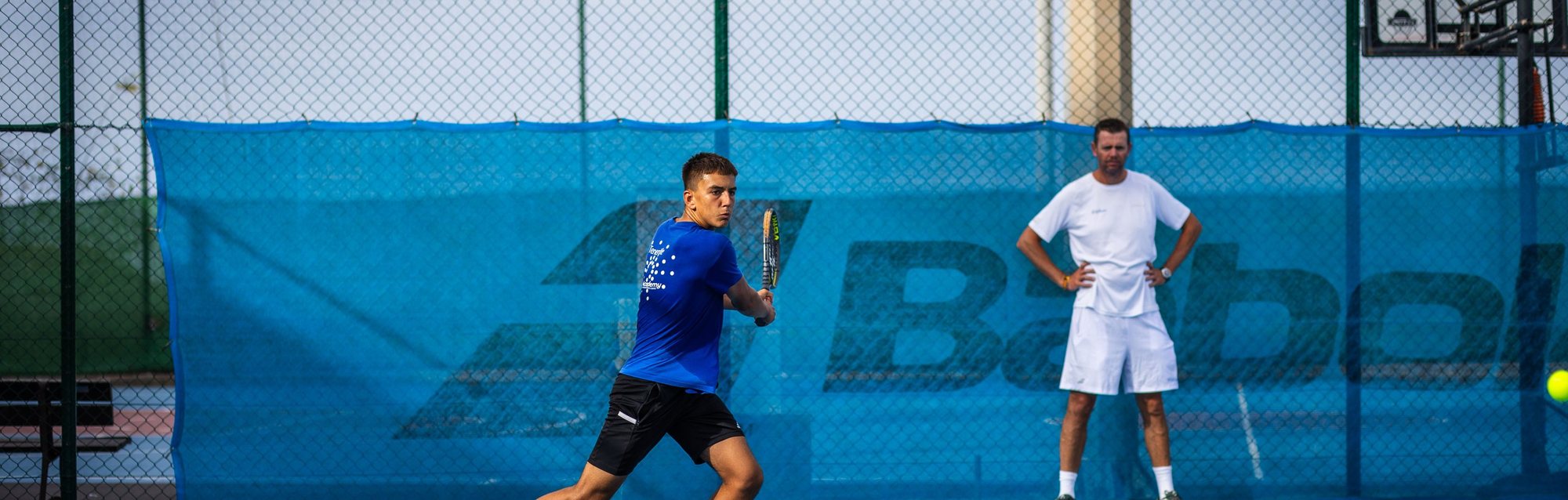 A player mid-rally with his coach watching from the baseline